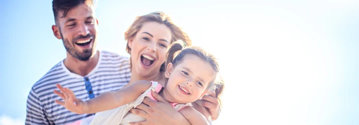 A group of three people outdoors under bright sunlight, with one person holding a child. They appear to be enjoying a casual moment together.